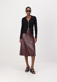 Cardigan noir en maille côtelée avec fermeture éclair, associé à une jupe midi en cuir bordeaux. Sandales à talons noires avec accents de boucles.