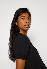Vestido preto com mangas bufantes, apresentando um corpete texturizado. O cabelo é longo e ondulado, com um fundo neutro que cria contraste.