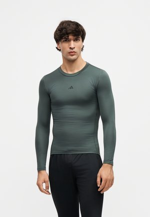 Homme portant un t-shirt de sport moulant à manches longues vert foncé et un pantalon noir, debout devant un fond clair uni.