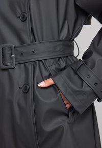 Black waterproof trench coat featuring a belted waist, large buttons, and pockets, made from smooth, glossy material with a tailored fit.