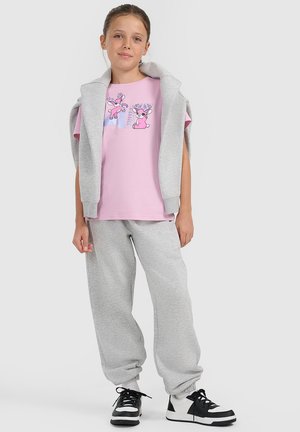 Dziewczynka ubrana w różowy t-shirt z dwoma kreskówkowymi jeleniami, szare spodnie dresowe, szarą bluzę przewieszoną przez ramiona oraz czarno-białe trampki.