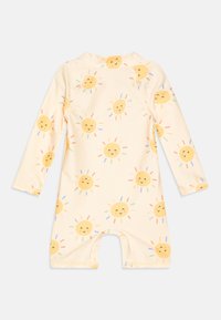 Sursuit à manches longues de couleur jaune clair, avec un motif répétitif de visages de soleil souriants entourés de rayons colorés. Conçu pour le confort et le jeu.
