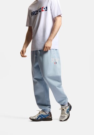 Osoba ubrana w białą koszulkę z nadrukiem, jasnoniebieskie dresy z napisem „Prosto Sport” oraz białe i niebieskie sportowe buty, stojąca na jednolitym tle.