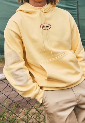 Personne portant un sweat à capuche jaune pâle avec le logo "air max", debout avec une main dans la poche, appuyée contre une clôture en grillage extérieur.