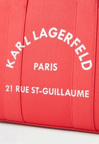 Raudona medžiaga su tekstūru rodo "Karl Lagerfeld Paris" ir "21 Rue St-Guillaume" baltomis paryškintomis raidėmis.