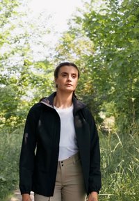 Veste noire imperméable avec capuche, dotée d'une fermeture éclair sur le devant et de poches latérales. Portée par-dessus une chemise blanche et un pantalon beige dans un décor verdoyant.