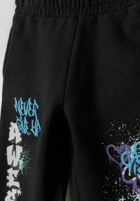 Zwarte trainingsbroek met een elastische tailleband, voorzien van kleurrijke graffiti-achtige tekst en spetters in blauwe en paarse accenten.
