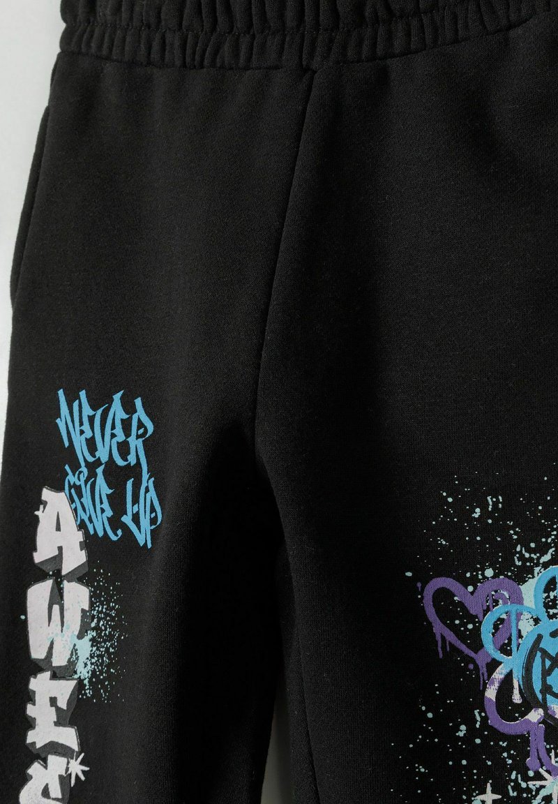 Pantalons de survêtement noirs avec une taille élastique, ornés de textes et de motifs éclaboussés de style graffiti en accents bleu et violet.