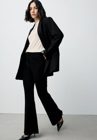 Black blazer over a beige ribbed top, paired with black flared trousers. The outfit features sleek textures and pointed patent leather heels.