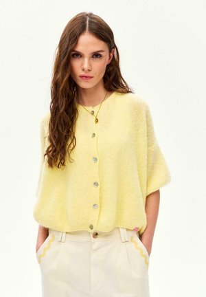 Młoda kobieta o długich brązowych włosach, ubrana w bladoróżowy kardigan zapinany na guziki i kremowe spodnie z wysokim stanem oraz falistym wykończeniem, z rękami w kieszeniach.