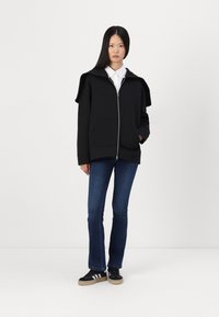 Hoodie noir zippé avec un design unique sur les épaules, associé à un jean évasé bleu et des baskets noires avec des rayures blanches.