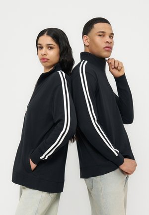 Du jauni suaugusieji stovi nugara į nugarą dėvėdami juodus striukes su užtrauktukais ir baltomis juostelėmis rankovėse bei šviesios spalvos kelnes, fone paprasta balta sienelė.