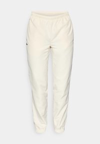 Pantaloni sportivi color crema con vita elastica, gambe aderenti e un piccolo logo verde scuro sul lato sinistro. Tessuto liscio e leggero.