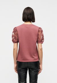 Γυναίκα που φοράει μπλούζα σε ζαχαρί ροζ με σύντομα μανίκια από δαντέλα με φουσκωτά λουλούδια και μαύρο παντελόνι από τεχνητό δέρμα, φαίνεται από πίσω.