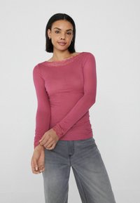 Langärmliges pinkes Oberteil mit Spitzenakzenten entlang des Boote-Ausschnitts und den Säumen, enganliegendes Design mit weicher Textur, kombiniert mit grauen Jeans.