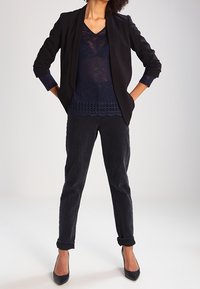 Svart blazer med notch-krage, lager på en marinblå topp med spetsdetaljer, ihop med svarta uppvikta byxor och svarta klackar.