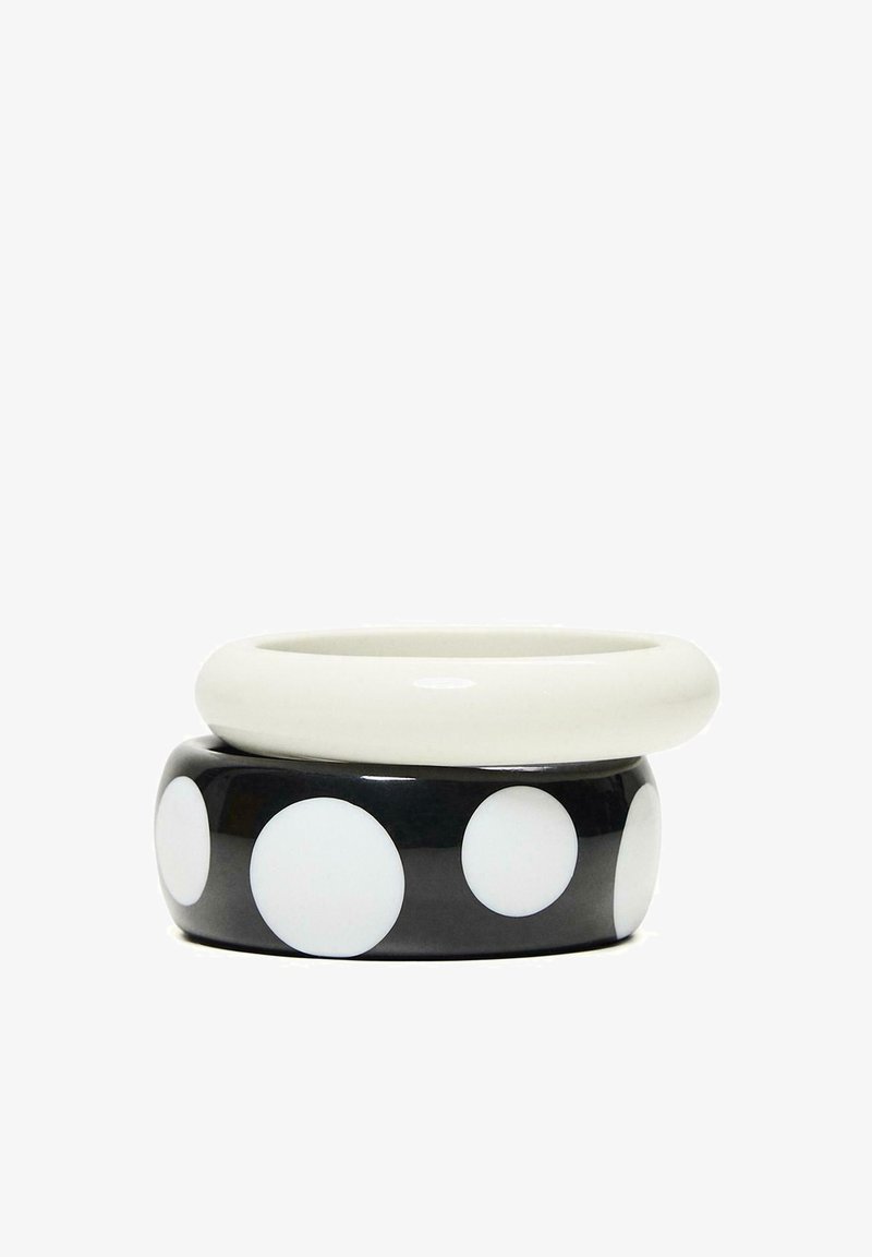 Deux bracelets empilés, un blanc uni et un noir à gros pois blancs, présentés sur un fond blanc.