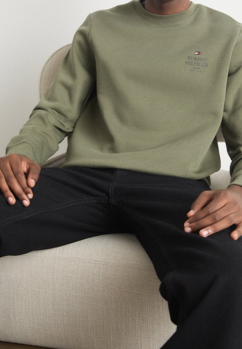 Persona seduta su una sedia beige, indossa un felpa olive verde di Tommy Hilfiger e pantaloni neri, con le mani appoggiate sulle cosce.