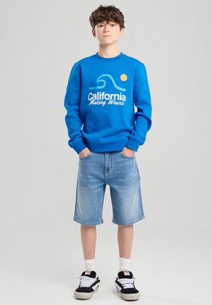 Sudadera azul con un gráfico que dice "California Making Waves", combinada con pantalones cortos de mezclilla claros y zapatillas negras con acentos blancos.