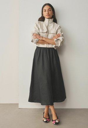 Frau mit langen dunklen Haaren steht mit verschränkten Armen, trägt eine beige Reißverschlussjacke, einen schwarzen Midi-Rock, goldene Creolen, ein Armband und florale Ballerinas.