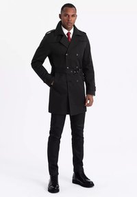 Czarny płaszcz trench z podwójnym zapięciem, paskiem w talii i epoletami na ramionach, noszony na białej koszuli i czerwonym krawacie, w połączeniu z czarnymi spodniami i butami.