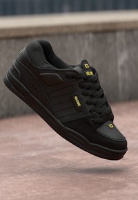 Czarne sneakersy o gładkiej skórzanej fakturze, z trzema poziomymi paskami, okrągłymi sznurówkami i żółtymi akcentami loga. Solidna gumowa podeszwa.