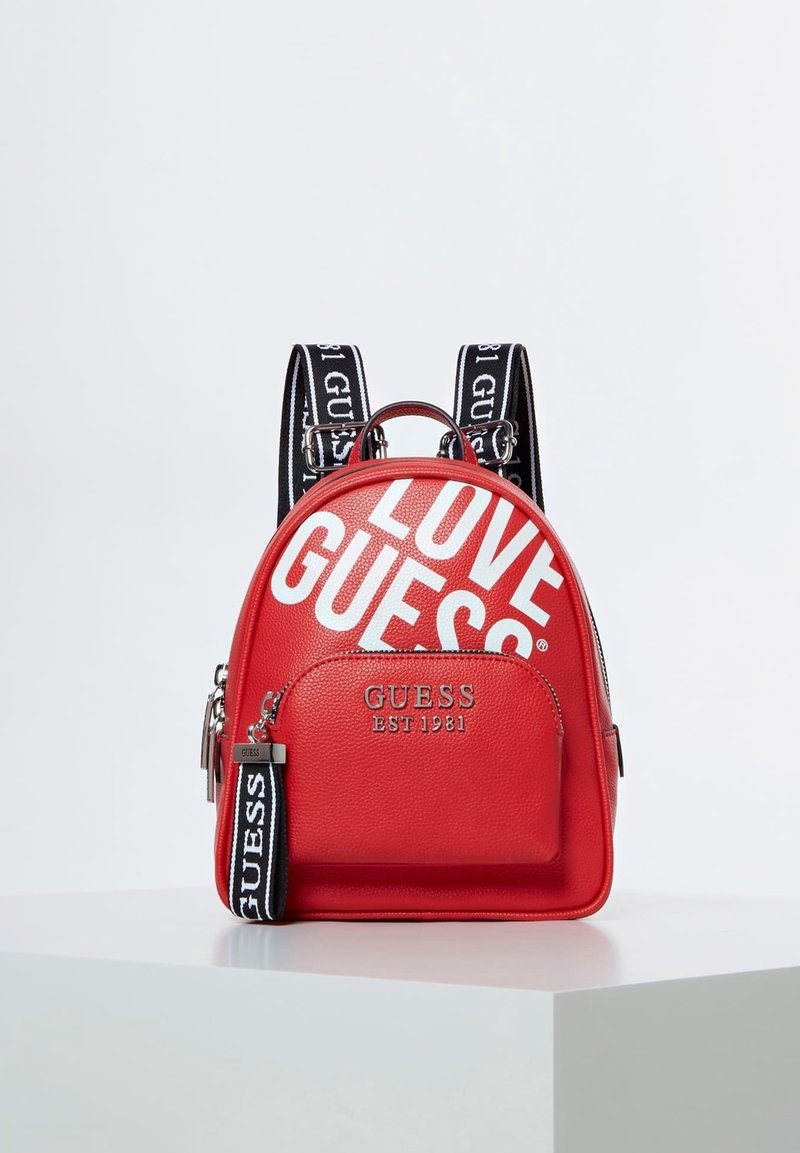 Mini sac à dos rouge Guess avec poche avant, bretelles avec logo noir et blanc, et grand texte blanc "LOVE GUESS" sur le compartiment principal.