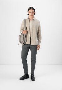 Camisa de botones color beige claro sobre una blusa crema, combinada con pantalones grises slim-fit y zapatos negros. Una bolsa gris cuelga del hombro.