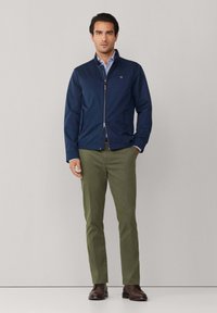 Mężczyzna ubrany w granatową kurtkę na zamek, niebieską koszulę w paski, oliwkowe spodnie i brązowe eleganckie buty, stojący na tle jasnego, jednolitego tła.