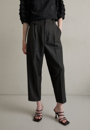 Persoon die zwarte geruite hoge taille broek en zwarte sandalen met bandjes en hoge hakken draagt, staand tegen een eenvoudige lichte muur.