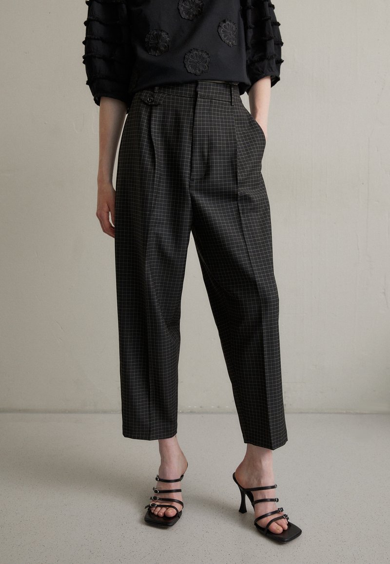Personne portant un pantalon à carreaux noirs taille haute et des sandales noires à talons hauts et brides, debout contre un mur clair uni.