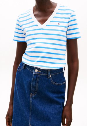 Osoba ubrana w biały t-shirt z niebieskimi paskami i małym logo na piersi, wpuszczony w ciemną, wysoką spódnicę z denimu.