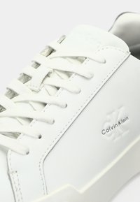 Λευκά δερμάτινα αθλητικά παπούτσια με επίπεδα λευκά κορδόνια, υφή στις πλευρές και ξεχωριστό τυπωμένο λογότυπο στην εξωτερική πλευρά. Μινιμαλιστικός σχεδιασμός με λεία επιφάνεια.