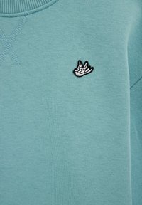 Svetlomodrá mikina z mäkkej tkaniny s malým vyšívaným čierno-bielym logom vtáka na ľavej strane hrudníka. Textúrovaný výstrih.