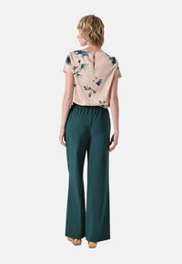 Beige blouse met bloemenpatroon en korte mouwen, met een keyhole back, gecombineerd met groene, wijde broek en gouden schoenen.