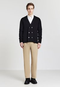 Junger Mann, der vor einem einfarbigen Hintergrund steht und eine schwarze doppelt knöpfbare Strickjacke, ein weißes Hemd, beige Hosen und schwarze Schuhe trägt.