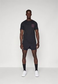 Černé atletické tričko s růžovým potiskem, doplněné tmavými šortkami a bílými teniskami. Model stojí na neutrálním pozadí.