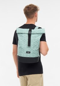 Joven con una mochila de patrón geométrico en verde menta con fondo y correa negra sobre un fondo blanco.