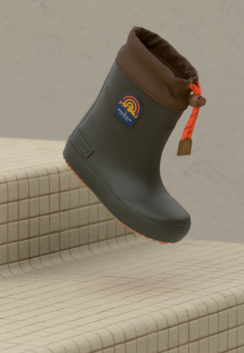 Botte d'hiver étanche vert foncé avec un bord en tissu marron, cordon orange et badge d'explorateur de montagne, flottant au-dessus de marches carrelées beiges.