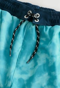 Shorts de bain dans un motif tie-dye bleu clair, avec une ceinture élastique marine, un cordon de serrage noir et une poche latérale.