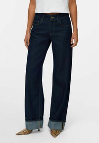 Jean en denim foncé à pattes larges avec un design à cinq poches, présentant des coutures contrastées et des ourlets retroussés. Associé à des chaussures à bout pointu à motifs.