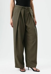 Olijfgroene broek met brede pijpen en plooien, met een tailleband met riemlussen en een gladde textuur. Gecombineerd met zwarte open-teenhakken.