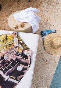 Shorts colorés sur une chaise longue blanche à côté d'une table ronde avec une serviette blanche et des lunettes de soleil, un chapeau de paille près du bord de la piscine sur un sol en pierre.
