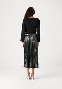 Top nero corto a maniche lunghe abbinato a una gonna plissettata verde scuro metallica, caratterizzata da pieghe orizzontali e una finitura liscia. Décolleté bianchi con tacco spesso.