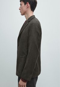 Blazer marrone a spina di pesce con rever a lancia, chiusura a due bottoni e tasche laterali. Indossato sopra a un maglione rosso testurizzato, mostrato di profilo.