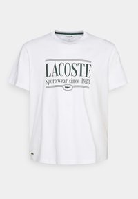 Bílý bavlněný tričko s zeleným textem, který zní "LACOSTE Sportswear od roku 1933" a malým logem Lacoste poblíž lemu. Krátké rukávy.