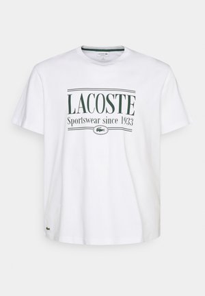 Camiseta blanca de manga corta con la impresión negra "LACOSTE", combinada con pantalones cargo beig y zapatillas deportivas grises con acentos negros.