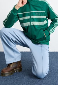 Grön zip-up jacka med ljusgröna ränder; ljusblå löst sittande byxor; bruna snörskor med tjocka sulor.