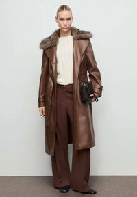 Manteau en cuir marron avec un col en fourrure, superposé à un pull beige et un pantalon large marron, accompagné d'un petit sac à main noir.