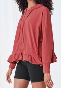 Haut à capuche zippé rouge avec un ourlet volanté, des manches longues et une capuche. Fabriqué en matériau doux. Associé à un short noir.